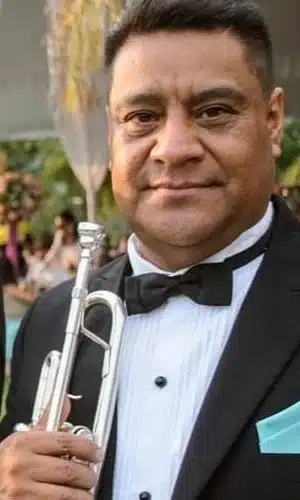 Trompetista Javier Bautista vestido de smoking, sosteniendo su trompeta mientras espera al inicio de la participación del grupo versátil Alfamusic