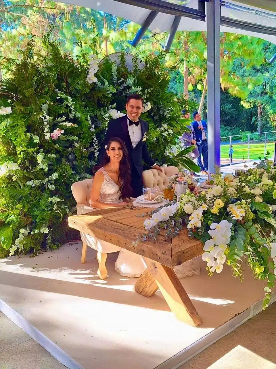 A bride and groom pose for a photo in front of a floral arrangement.